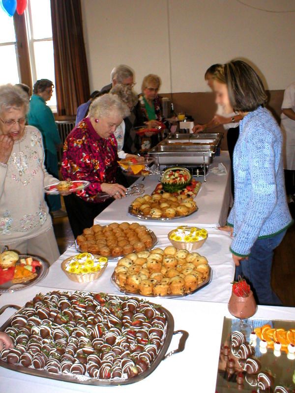 People walk along buffet table full of muffins, fruit, chocolate covered strawberries and more.